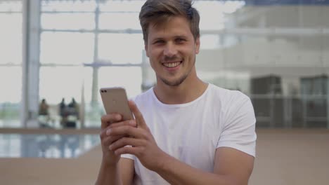Smiling-young-man-holding-smartphone-and-looking-at-camera.